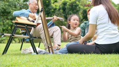 Mutlu Asyalı nesiller parkta pikniğin tadını çıkartıyor büyükbabam gitar çalıyor neşeli bir açık hava hayatı yaratıyor.