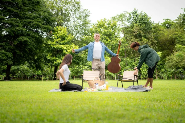 Mutlu Asyalı nesiller parkta pikniğin tadını çıkartıyor büyükbabam gitar çalıyor neşeli bir açık hava hayatı yaratıyor.