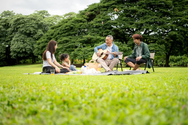 Mutlu Asyalı nesiller parkta pikniğin tadını çıkartıyor büyükbabam gitar çalıyor neşeli bir açık hava hayatı yaratıyor.