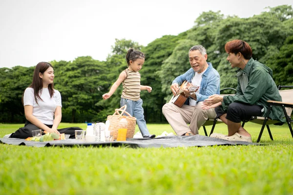 Mutlu Asyalı nesiller parkta pikniğin tadını çıkartıyor büyükbabam gitar çalıyor neşeli bir açık hava hayatı yaratıyor.