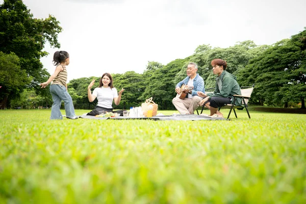 Mutlu Asyalı nesiller parkta pikniğin tadını çıkartıyor büyükbabam gitar çalıyor neşeli bir açık hava hayatı yaratıyor.