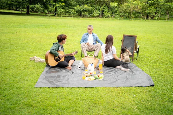 Mutlu Asyalı nesiller parkta pikniğin tadını çıkartıyor büyükbabam gitar çalıyor neşeli bir açık hava hayatı yaratıyor.