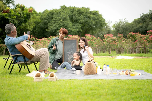 Mutlu Asyalı nesiller parkta pikniğin tadını çıkartıyor büyükbabam gitar çalıyor neşeli bir açık hava hayatı yaratıyor.