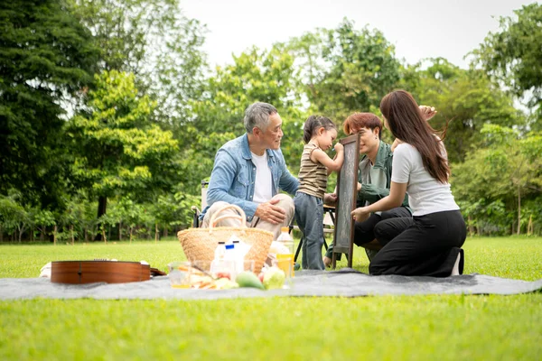 Mutlu Asyalı nesiller parkta pikniğin tadını çıkartıyor büyükbabam gitar çalıyor neşeli bir açık hava hayatı yaratıyor.