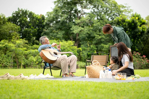 Mutlu Asyalı nesiller parkta pikniğin tadını çıkartıyor büyükbabam gitar çalıyor neşeli bir açık hava hayatı yaratıyor.