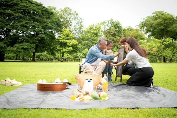 Mutlu Asyalı nesiller parkta pikniğin tadını çıkartıyor büyükbabam gitar çalıyor neşeli bir açık hava hayatı yaratıyor.