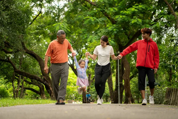 Asyalı bir aile parkta birlikte koşuyor. Ebeveynler, çocuklar ve büyükbabalar dışarıda egzersiz yapıyor, sağlıklı yaşam tarzını ve aile bağlarını destekliyor.