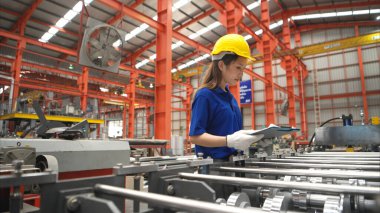 Genç bayan işçi, güvenlik kaskları ve diğer ekipmanlarla üretim hattındaki makineleri denetliyor.
