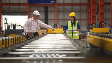 Kıdemli mühendisler ve genç bayan işçiler güvenlik miğferleri ve diğer ekipmanlarla üretim hattındaki makineleri konuşuyor ve denetliyorlar.