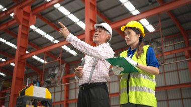 Kıdemli mühendisler ve genç bayan işçiler güvenlik miğferleri ve diğer ekipmanlarla üretim hattındaki makineleri konuşuyor ve denetliyorlar.