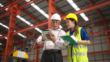 Kıdemli mühendisler ve genç bayan işçiler güvenlik miğferleri ve diğer ekipmanlarla üretim hattındaki makineleri konuşuyor ve denetliyorlar.