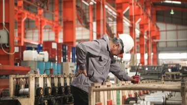 Fabrika makineleri üzerinde çalışırken sırt ağrısı çeken koruyucu ekipmanlı sanayi işçisi.