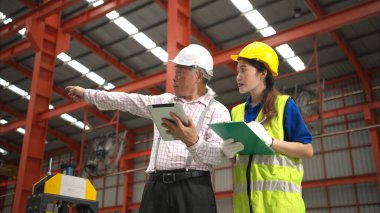 Kıdemli mühendisler ve genç bayan işçiler güvenlik miğferleri ve diğer ekipmanlarla üretim hattındaki makineleri konuşuyor ve denetliyorlar.