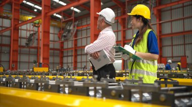 Kıdemli mühendisler ve genç bayan işçiler güvenlik miğferleri ve diğer ekipmanlarla üretim hattındaki makineleri konuşuyor ve denetliyorlar.