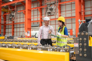 Kıdemli mühendisler ve genç bayan işçiler güvenlik miğferleri ve diğer ekipmanlarla üretim hattındaki makineleri konuşuyor ve denetliyorlar.