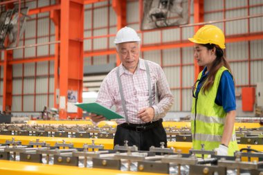 Kıdemli mühendisler ve genç bayan işçiler güvenlik miğferleri ve diğer ekipmanlarla üretim hattındaki makineleri konuşuyor ve denetliyorlar.
