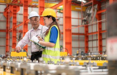 Kıdemli mühendisler ve genç bayan işçiler güvenlik miğferleri ve diğer ekipmanlarla üretim hattındaki makineleri konuşuyor ve denetliyorlar.