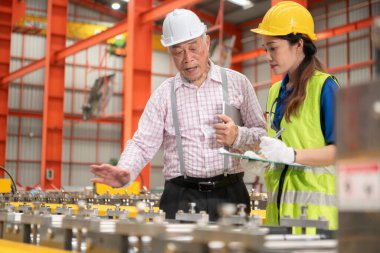 Kıdemli mühendisler ve genç bayan işçiler güvenlik miğferleri ve diğer ekipmanlarla üretim hattındaki makineleri konuşuyor ve denetliyorlar.
