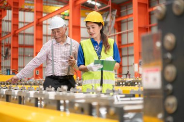 Kıdemli mühendisler ve genç bayan işçiler güvenlik miğferleri ve diğer ekipmanlarla üretim hattındaki makineleri konuşuyor ve denetliyorlar.
