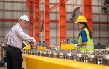 Kıdemli mühendisler ve genç bayan işçiler güvenlik miğferleri ve diğer ekipmanlarla üretim hattındaki makineleri konuşuyor ve denetliyorlar.