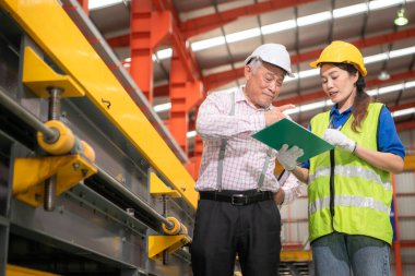 Kıdemli mühendisler ve genç bayan işçiler güvenlik miğferleri ve diğer ekipmanlarla üretim hattındaki makineleri konuşuyor ve denetliyorlar.