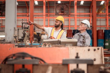 Kıdemli mühendisler ve genç işçiler güvenlik miğferleri ve diğer ekipmanlarla üretim hattındaki makineleri konuşuyorlar ve denetliyorlar.