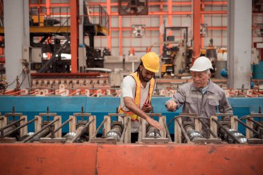 Kıdemli mühendisler ve genç işçiler güvenlik miğferleri ve diğer ekipmanlarla üretim hattındaki makineleri konuşuyorlar ve denetliyorlar.