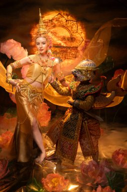 Geleneksel Tayland dansı gösterisinde zarif bir bayan dansçı ve altın kostümlü maskeli bir Khon sanatçısı, zarafet ve kültür mirasını sembolize ediyor.