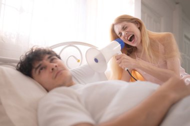 Sabahları ortağını uyandırmak için megafon kullanan oyuncu kadın, yatakta kulaklarını kapatıyor.