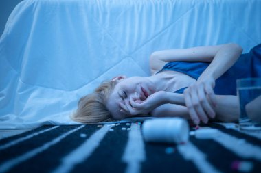 Depresyonda olan genç bir kadın yere düşmüş haplarla uzanıyor, zihinsel sağlık sorunları, bağımlılık ve duygusal çöküntüleri tasvir ediyor.