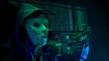 Maske takan ve kapüşonlu bir bilgisayar korsanı karanlık bir odada dijital verilerle etkileşime geçerek siber suç ve online güvenlik tehditlerini temsil ediyor.