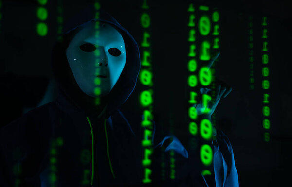 Анонимный хакер в маске и толстовке, взаимодействующий с цифровыми данными в темной комнате, представляющий угрозу киберпреступности и безопасности в Интернете