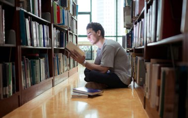 Kütüphane zemininde oturan genç bir adam konsantrasyonlu kitap okuyor.