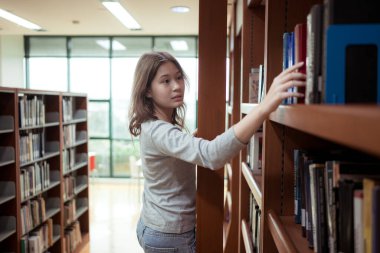 Genç bir kadın sessiz bir kütüphane koridorundaki kitaplığın en üst rafından bir kitap almak için merdiven kullanıyor.