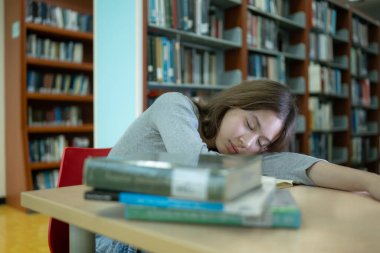 Genç bir kız kütüphanenin masasında oturuyor, kitap için bilgi arıyor ve okuyor.