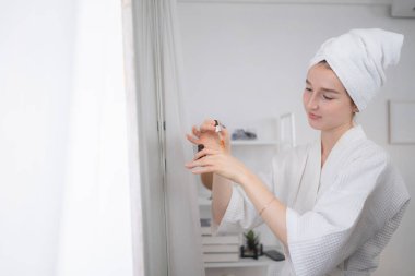 Genç bir kadın aynanın karşısında duruyor, cilt bakım serumu uygulayarak yüzüne bakıyor.