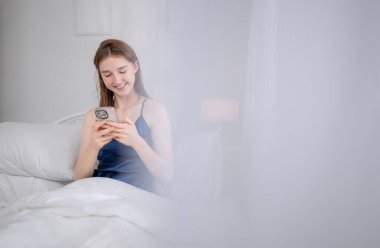 Genç bir kadın yatakta oturuyor ve sabah yatağında akıllı telefonunu kontrol ediyor.