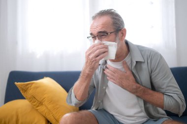 Kanepede oturan yaşlı bir adam öksürüyor, hapşırıyor ve grip virüsü belirtileri gösteriyor.
