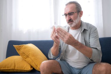 Kanepede oturan yaşlı bir adam öksürüyor, hapşırıyor ve grip virüsü belirtileri gösteriyor.