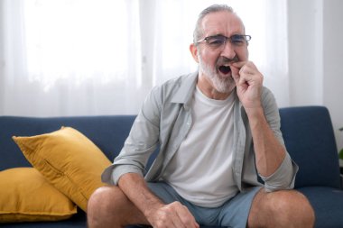 Kanepede oturan yaşlı bir adam diş çürüğünün ya da ağız hastalığının semptomlarını gösteren acı içinde dişini tutuyor.