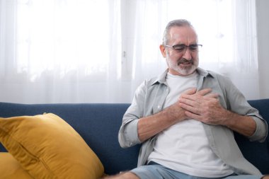 Kanepede oturan ve göğsünü ağrı içinde tutan yaşlı bir adam kalp krizi, göğüs ağrısı ya da evdeki acil durumu gösteriyor.