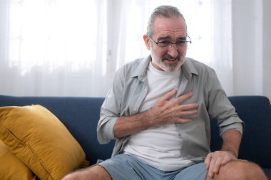 Kanepede oturan ve göğsünü ağrı içinde tutan yaşlı bir adam kalp krizi, göğüs ağrısı ya da evdeki acil durumu gösteriyor.