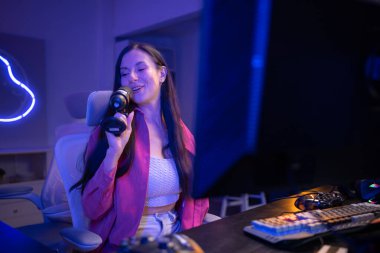 Bilgisayar ekranı ile etkileşime girerken mikrofona şarkı söyleyen bir içerik yaratıcısı ya da flaması. Neon ışıklı oda ve oyun ekipmanları canlı ve yaratıcı bir akış ortamı yaratır.