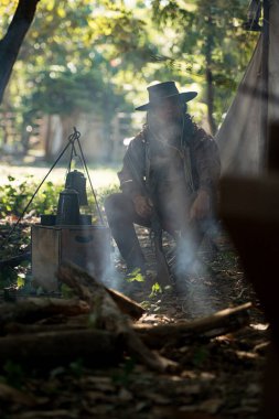 Dumanlı bir orman kampında sakince oturan yaşlı bir kovboy elinde tüfekle ağaçların arasından güneş ışınları geçerken...