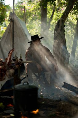Dumanlı bir orman kampında sakince oturan yaşlı bir kovboy elinde tüfekle ağaçların arasından güneş ışınları geçerken...