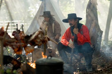 Bir grup kovboy kamp ateşinde yemek hazırlıyor, sıcak ve kırsal alanda birlikte et kızartıyorlar.