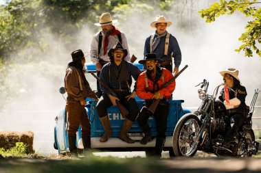 Bir grup kovboy, duman ve güneş ışığıyla çevrili eski bir pikap ve motosiklet etrafında birlikte gülüyor.