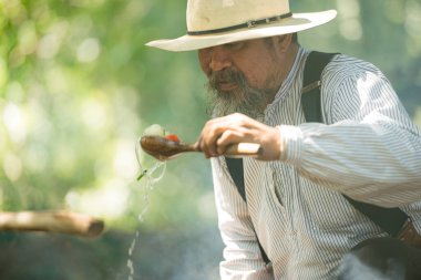Kırsal bir kamp ateşinde yemek hazırlayan bir kovboy, yanında et pişirirken buharı tüten bir çorba tenceresini tatlandırıyor ve karıştırıyor.