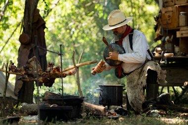 Kırsal bir kamp ateşinde yemek hazırlayan bir kovboy, yanında et pişirirken buharı tüten bir çorba tenceresini tatlandırıyor ve karıştırıyor.