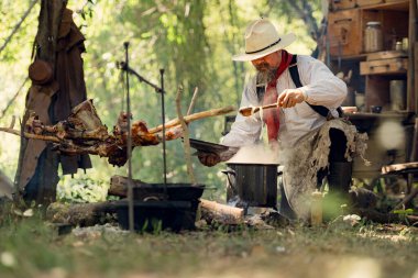 Kırsal bir kamp ateşinde yemek hazırlayan bir kovboy, yanında et pişirirken buharı tüten bir çorba tenceresini tatlandırıyor ve karıştırıyor.